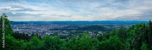 Wide panoramic view of Linz, Austria, framed by lush green forests. The cityscape spreads across the valley under a dramatic, cloudy sky, with the distant, faint silhouette of the Alps on the horizon.