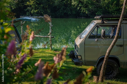 Vintage camper vans parked beside the Kolpa River on a summer day. Framed by flowers and trees, the campsite reflects a relaxed outdoor lifestyle