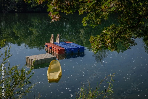Peaceful scene on the Kolpa River featuring a floating dock, pedal boat platform, and calm reflective water surrounded by lush green trees. Sunlight filters through the foliage, 
