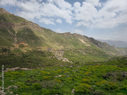 A mountain village nestled at the base of green slopes, surrounded by wildflowers and lush spring vegetation under a partly cloudy sky.
📍 Kamalla village, Hawraman Takht, Kurdistan