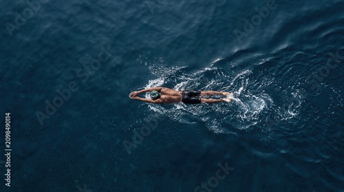 Aerial bird's-eye view of a male athlete swimming in deep blue open ocean water wearing a green swim cap and black trunks, creating white splashes and water ripples during training.