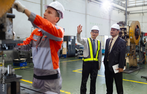 Engineers in hard hats inspect machinery on a factory floor while a technician operates equipment, emphasizing industrial production, safety practices, and supervision.