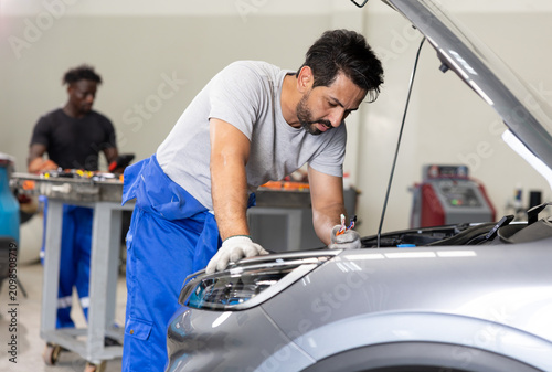Auto mechanic in blue overalls servicing a car engine under an open hood in a modern workshop, with tools and another technician working in the background.