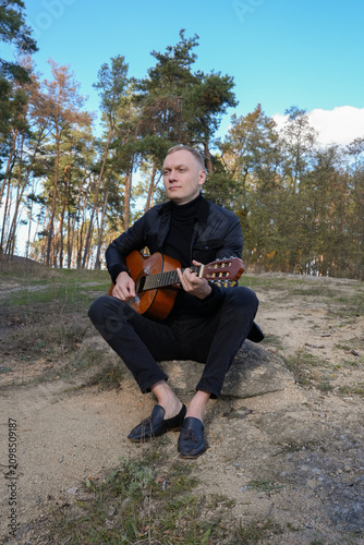 Beautiful blonde man playing acoustic guitar and singing relaxing enjoying outdoors on sunny autumn day. Artist learning and practicing playing musical instrument. Fall leaves autumnal weather 