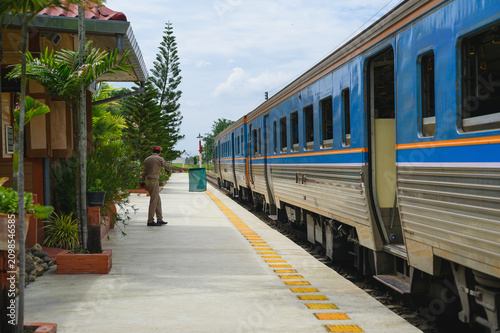Traditional thai train is pulling into rural station with person standing and waving green flag to signal safety, passenger train approaching platform. atmosphere of northern thailand rail travel