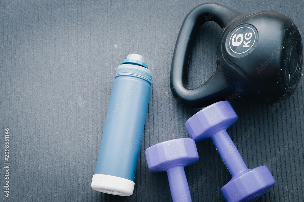 Fototapeta premium Top view flat lay of fitness equipment featuring a blue water bottle, two purple dumbbells, and a black 6kg kettlebell on a grey textured gym mat with copy space for text.