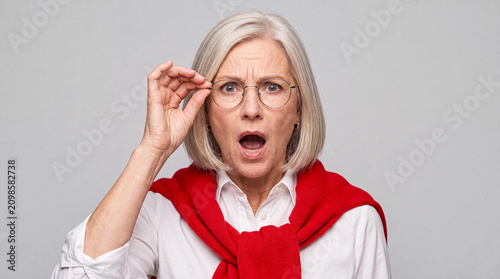 Fotografie Senior woman with glasses shows genuine surprise and shock, with mouth agape, on a light gray background
