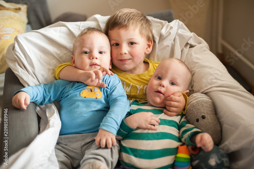 An older boy tenderly holds his two baby brothers close, nestled together in a soft armchair. Their sweet faces and innocent eyes radiate pure childhood joy in this heartwarming moment