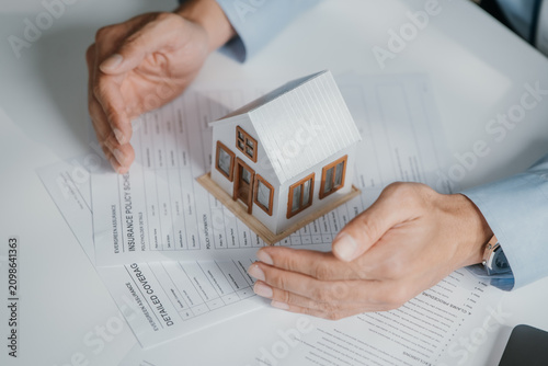Real estate agent or insurance broker hands protecting a model house on top of an insurance policy coverage document, representing property protection, risk management and secure investment.