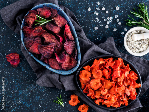 Baked beet and carrot slices with coarse sea salt scented rosemary. Vegan diet food idea and recipe. Healthy homemade beetroot and carrot chips in plate and ingredients on dark blue background. Copy