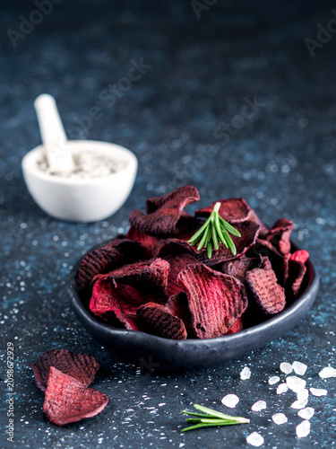 Homemade crispy vegan beet slices snack in a black bowl. Dark blue textured background with coarse sea salt, rosemary sprigs and copy space. Perfect for healthy diet and organic food marketing