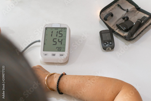 Foto Asian Woman Monitoring Vital Signs with Blood Pressure and Glucose Meters at Hom