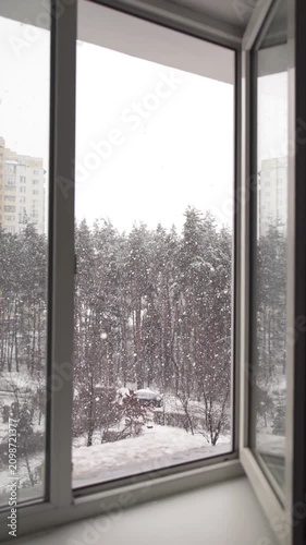 Looking through an open window at a heavy snowfall over a winter park.