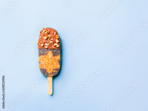 Healthy homemade plant-based chia seeds popsicle with raw carrot cake layer and chocolate coating with nuts. Minimalist flat lay on blue background. Chia popsicle with chocolate, copy space