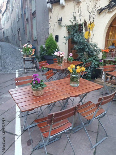 Bright floral arrangements on wooden bistro tables create a cozy outdoor cafe atmosphere in a charming courtyard, featuring roses, tulips and potted plants for a welcoming, romantic urban setting.