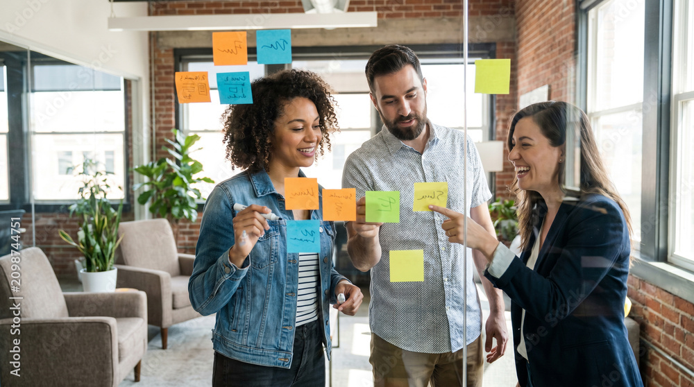 Obraz premium Creative professionals collaborate around glass whiteboard with colorful sticky notes, brainstorming ideas during startup meeting in modern office with natural daylight and copy space.