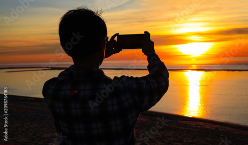 Back view with Silhouette of a man capturing a golden sunset reflection on the beach using a mobile phone a stunning ocean sunset during vacation