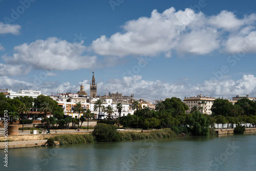 Fotografie Panoramic view of Seville across the Guadalquivir River, with the Giralda, historic architecture, palm trees and the Andalusian city skyline under a dramatic blue sky