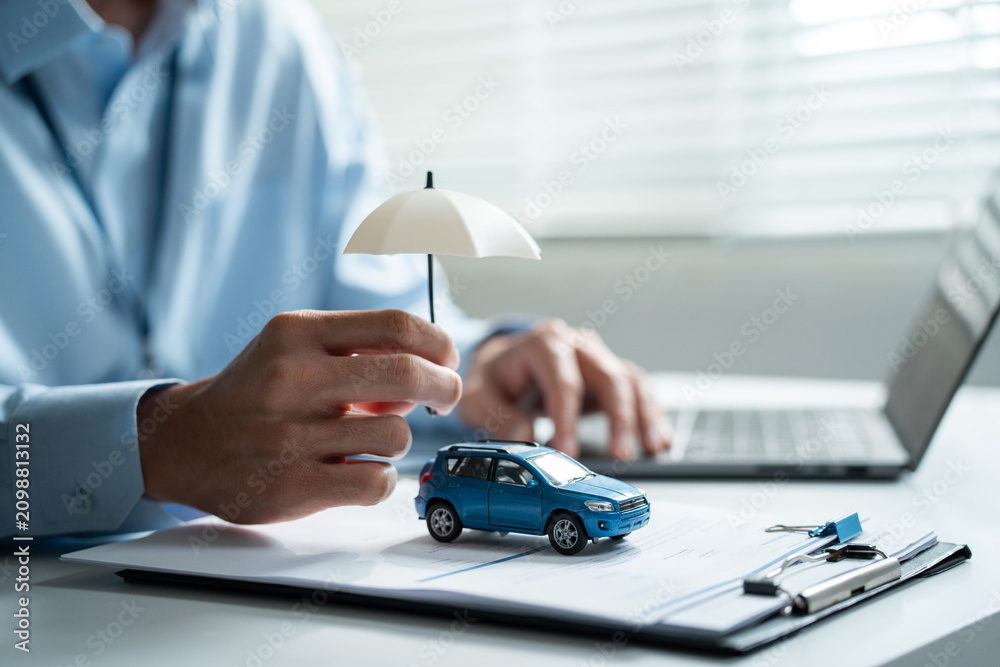Fototapeta premium Insurance agent holding umbrella over a blue toy car while working on a laptop computer, focusing on vehicle protection, digital finance, and online safety