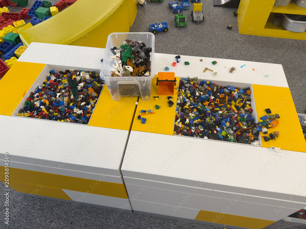 Fototapeta premium Lego bricks sorted in plastic bins on a table in a playroom, with colorful toy pieces and a children’s play area in the background