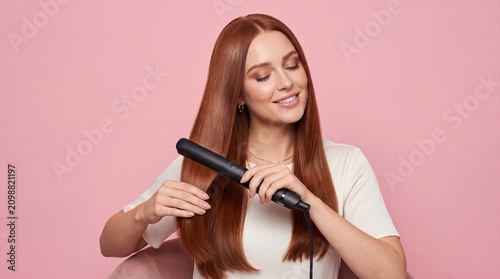 Young woman using hair straightener. Female styling long red hair with flat iron tool.