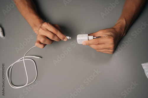 Close-up top view of man connecting USB cable to electronic cigarette charger on gray desk, showcasing charging process of vaping device. Concept of modern alternative to traditional smoking.