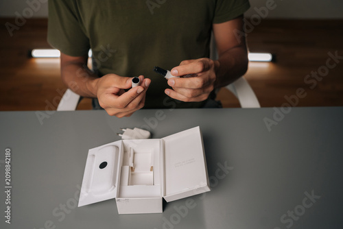 Close-up of smoker male sitting at table unpacking new electronic cigarette from box, holding heating element and cover. Concept of modern alternative to traditional smoking.