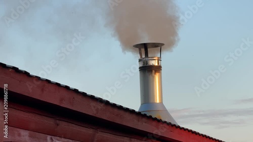 Smoke rises from a metal chimney on a red wooden roof against a cloudy sky. heating, winter home, cozy rural life.