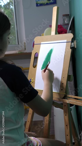 Little girl painting a green tree on an easel in a sunlit playroom
