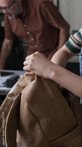 Vertical video of teenage girl unpacking notebooks from backpack on desk while middle aged teacher entering classroom and working on laptop. Perfect for back to school or learning concepts