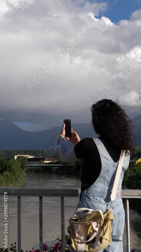 Woman photographing river and mountains on phone from a bridge in summer