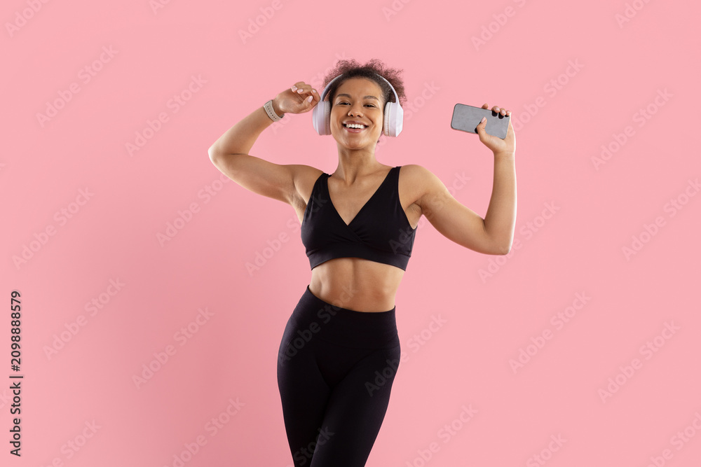 Fototapeta premium Happy young Black woman in fitness wear dancing with wireless headphones and smartphone, enjoying music against bright pink studio background with generous copy space