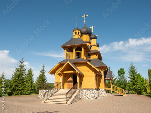 Картината върху платно A beautiful traditional Russian Orthodox church crafted from yellow timber logs features dark shingled roofs and multiple black onion domes topped with golden crosses under a clear blue sky