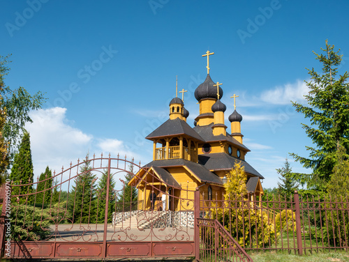 Φωτογραφία A beautiful traditional wooden Orthodox church with multiple black onion domes and golden crosses is seen behind a decorative brown metal fence and gate under a bright blue sky