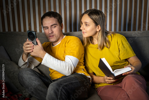 Woman hiding behind a book, secretly peeking at her partner's smartphone screen. Suspicion, jealousy, relationship control, and privacy invasion.