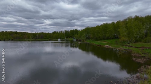 A high-altitude flight with a horizontal camera pan above a wide lake surrounded by forest on a cloudy summer day. Lake Bykovskoye, Novosibirsk Oblast, Russia.