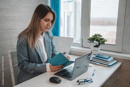 pretty young girl sit desktop pc hold pen write notepad shirt in home office indoors. A businesswoman makes notes in a notebook while sitting at her laptop. the student studies remotely.