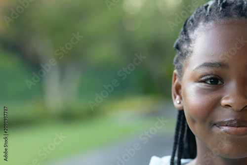 Child female smiling, showing braided hair and stud earring, wearing white top in park, copy space