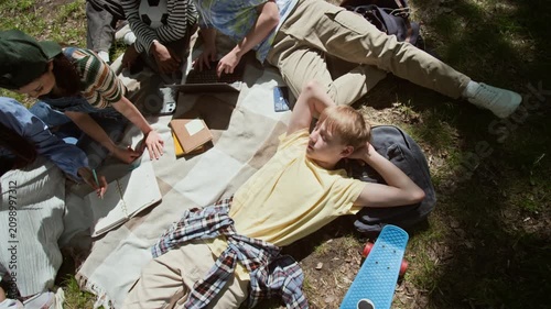 High angle shot of multiethnic teenagers studying together in park while Caucasian teenage boy lying on blanket relaxing. Footage for educational marketing and student lifestyle campaigns