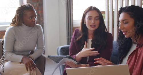 Diverse female coworkers reviewing laptop, tablet, exchanging printed pages in office aligning plan