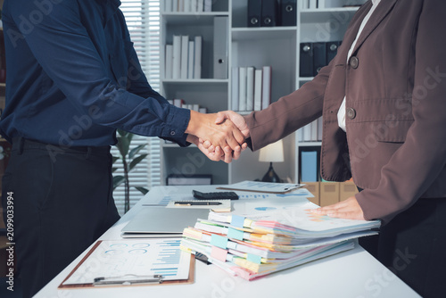 Successful Business Partnership Handshake: Two business people seal a deal with a firm handshake, signifying trust and collaboration in a modern office setting with stacks of documents on the desk.
