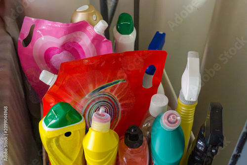 Colorful cleaning bottles stand in a cluttered storage area. Various shapes and bright hues dominate the visual. Some bottles have spray nozzles or caps visible