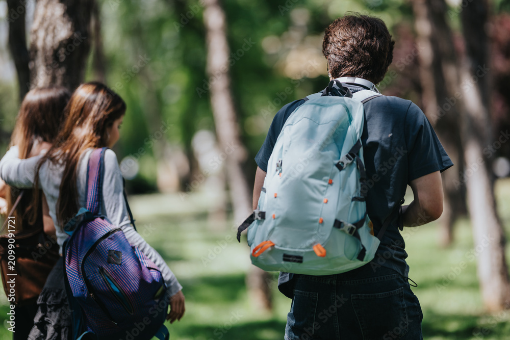 Naklejka premium Three students wearing backpacks walk along a shaded park path. Rear view shows them strolling together through green trees on a warm day.