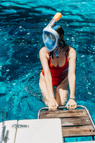 Woman in a red swimsuit and snorkel mask climbs onto the wooden swim platform of a boat. Boat charter leisure and snorkeling excursions for summer sea travel and watersport rental