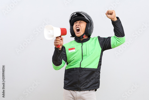 Portrait of an Asian male online motorbike taxi driver in a green jacket and helmet, standing holding a megaphone and shouting while raising his clenched fists, isolated on a gray background.