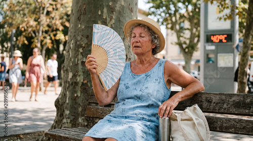 sweaty senior woman cooling herself with hand fan on bench during hot 40°C summer heatwave