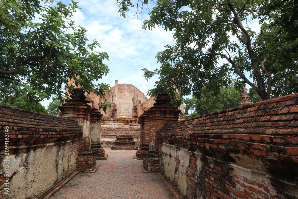Fototapeta premium Ancient Temple Path: A stone pathway winds through ancient brick walls toward a weathered temple ruin, framed by lush green trees under a cloudy sky.