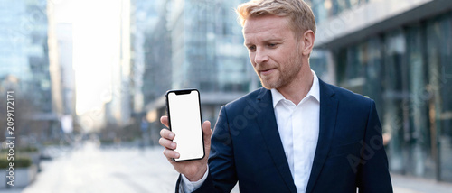 Ejecutivo de negocios sosteniendo un smartphone con pantalla blanca para mockup en un distrito financiero exterior. Concepto de tecnología móvil, finanzas y comunicación empresarial.