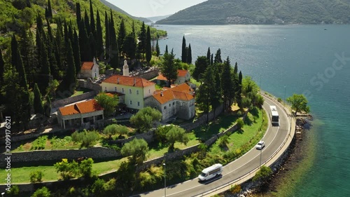 aerial view of a historic stone estate with orange tile roofs, a small bell tower and cypress trees above a curving coastal road on the shore of the bay of kotor, montenegro