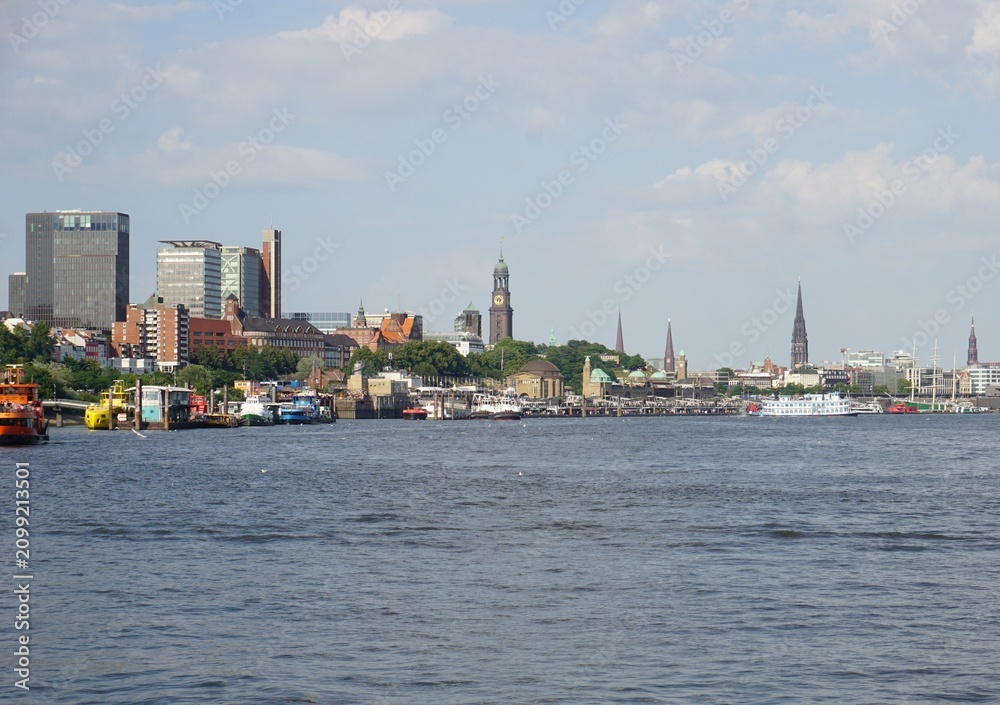 Fototapeta premium The skyline of Hamburg, Germany, viewed from river Elbe with various landmarks of the city, including the port of Hamburg, the St. Pauli landing bridges, and the bell towers of the main churches 